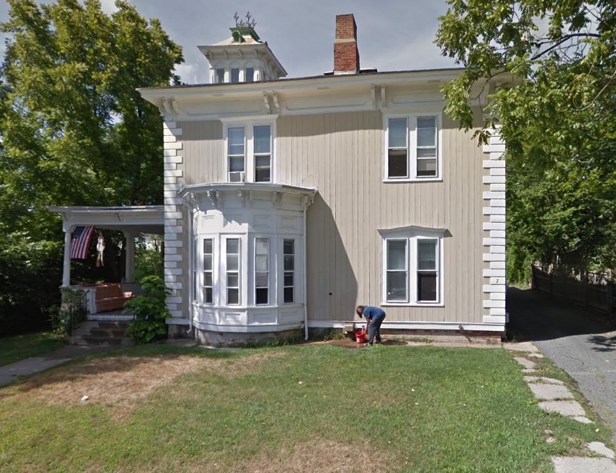 A person works on a red lawnmower in front of a two-story beige house with white trim, a bay window, and an American flag hanging on a porch to the left. Trees and a lawn surround the house.