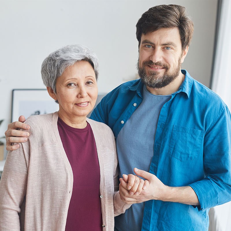 A middle-aged man and an older woman stand indoors, smiling, with the man’s arm around the woman and their hands clasped together.