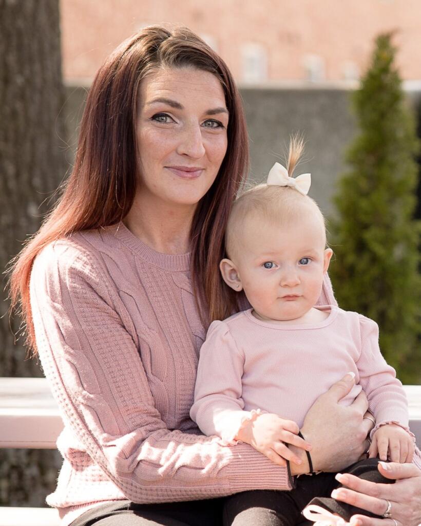 Michelle, a woman with long brown hair wearing a pink sweater sits on a bench outdoors, holding a baby girl in a matching pink outfit with a bow in her hair. Both are looking at the camera and smiling slightly.