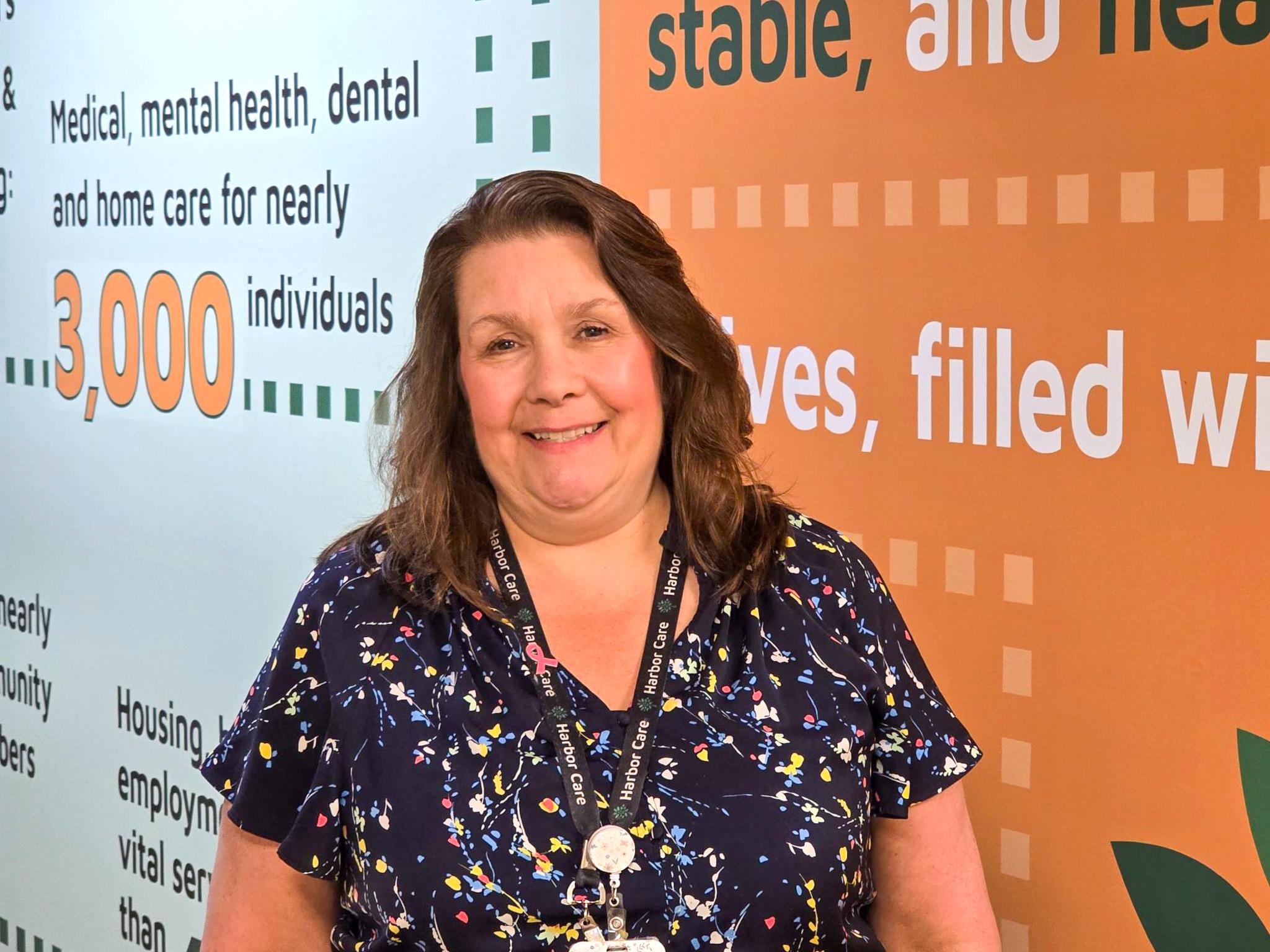 Pamela Urick A woman with brown hair and a navy patterned shirt stands in front of an orange and teal wall with text about medical, dental, and home care services.