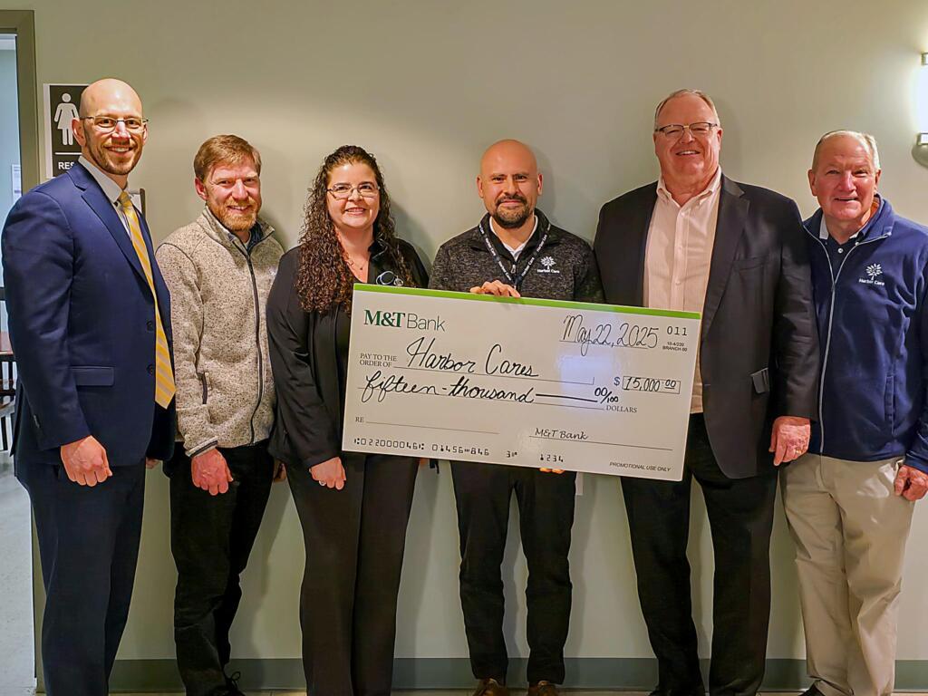 Six people stand indoors holding a large ceremonial check from M&T Bank to Harbor Corps for $15,000, dated May 22, 2025.