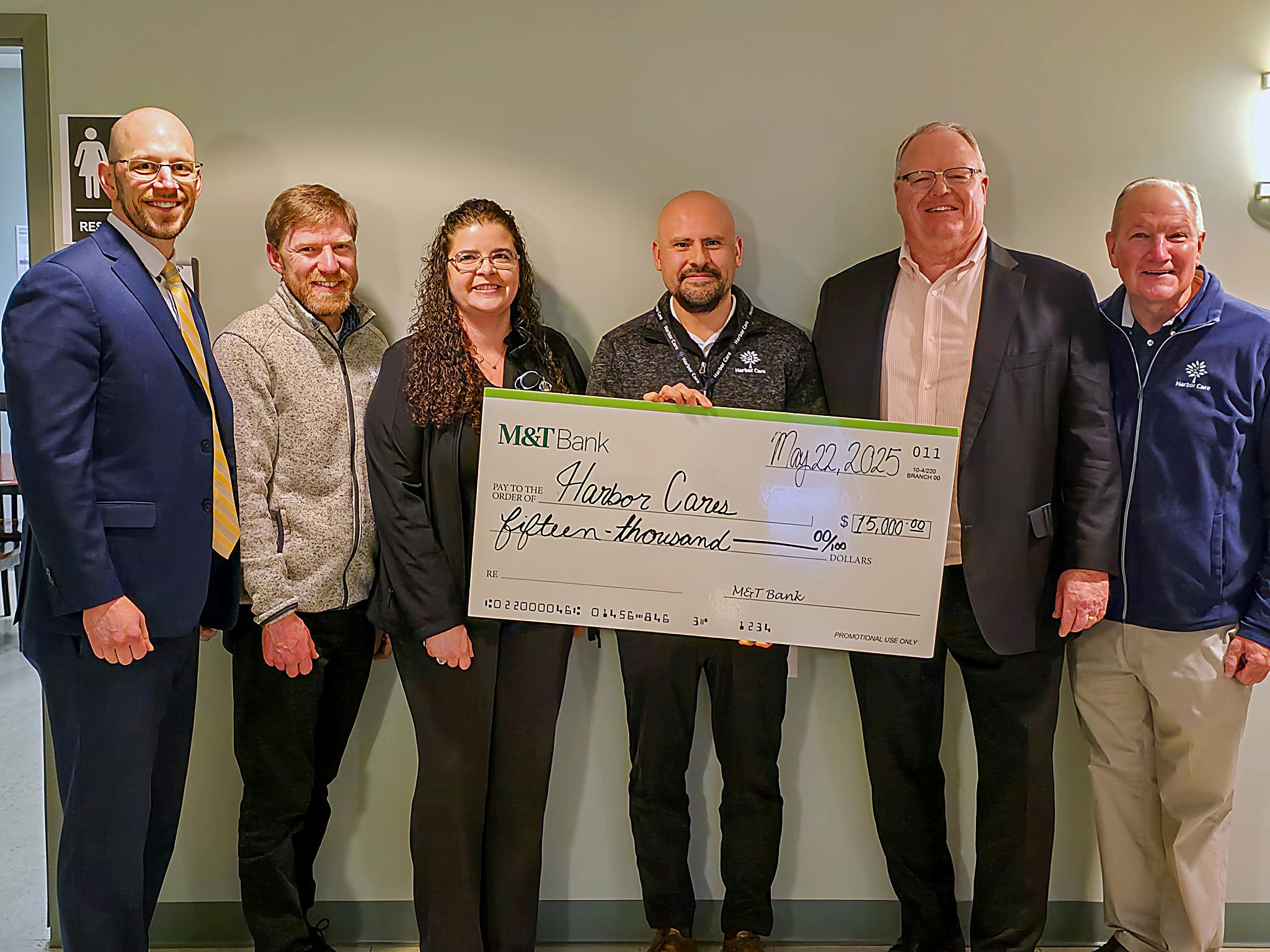 check_presentation_enhanced Six people stand indoors holding a large ceremonial check from M&T Bank to Harbor Corps for $15,000, dated May 22, 2025.
