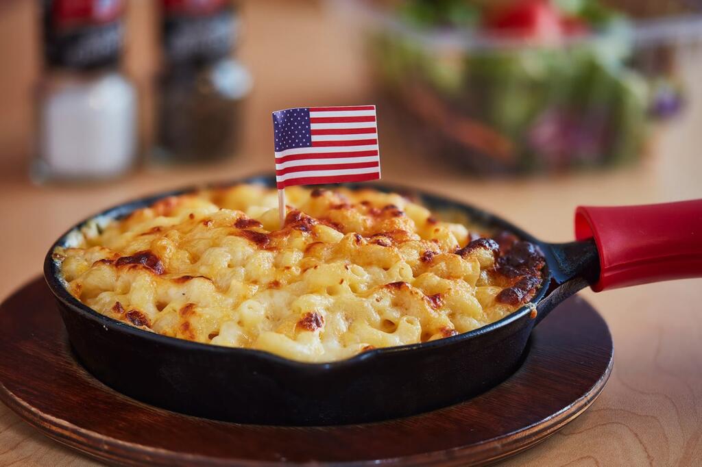 A skillet of baked macaroni and cheese topped with a small American flag, sitting on a wooden board, with salt, pepper, and a salad blurred in the background.