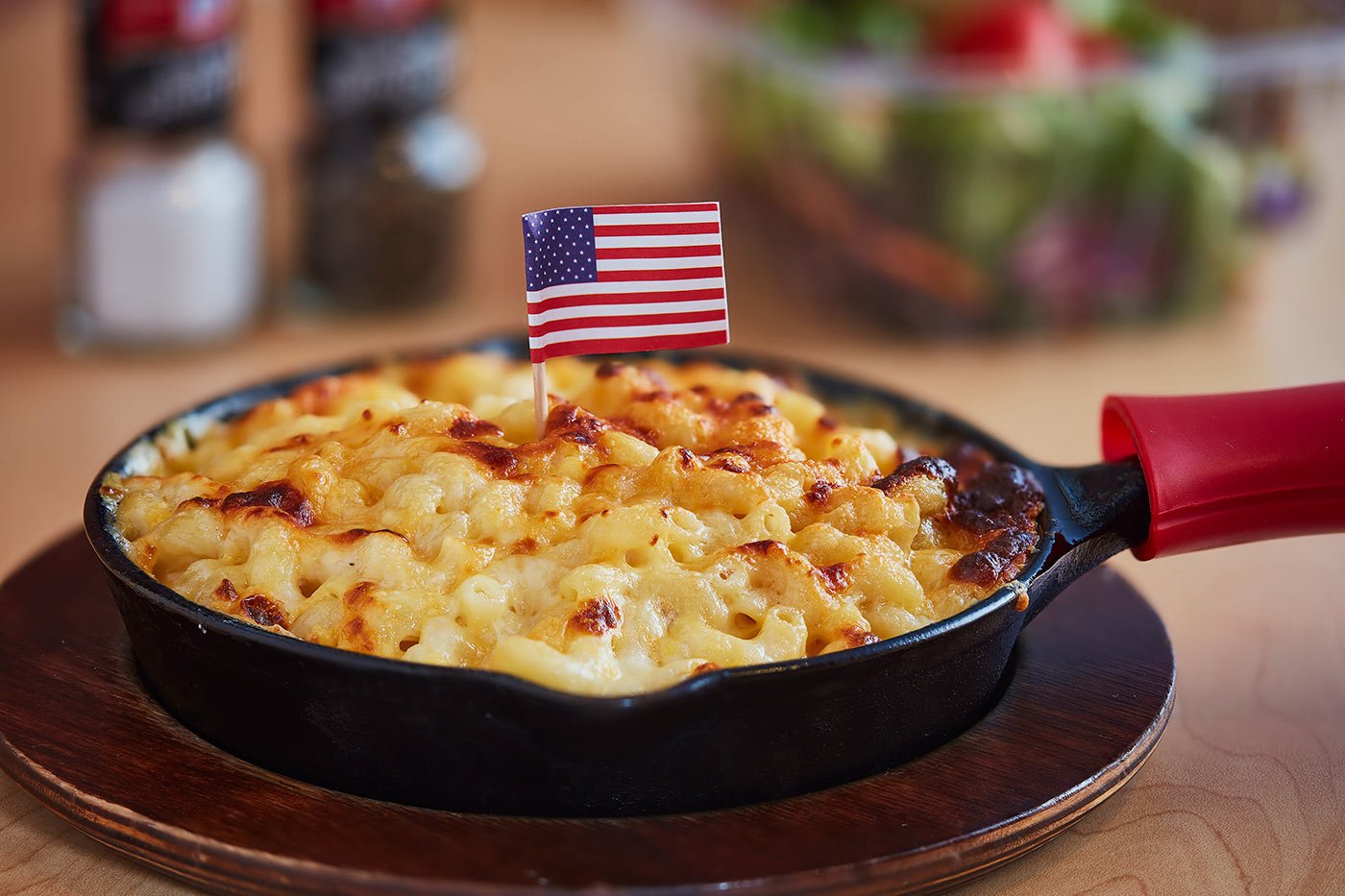 classic-american+mac+cheese+with+flag A skillet of baked macaroni and cheese topped with a small American flag, sitting on a wooden board, with salt, pepper, and a salad blurred in the background.
