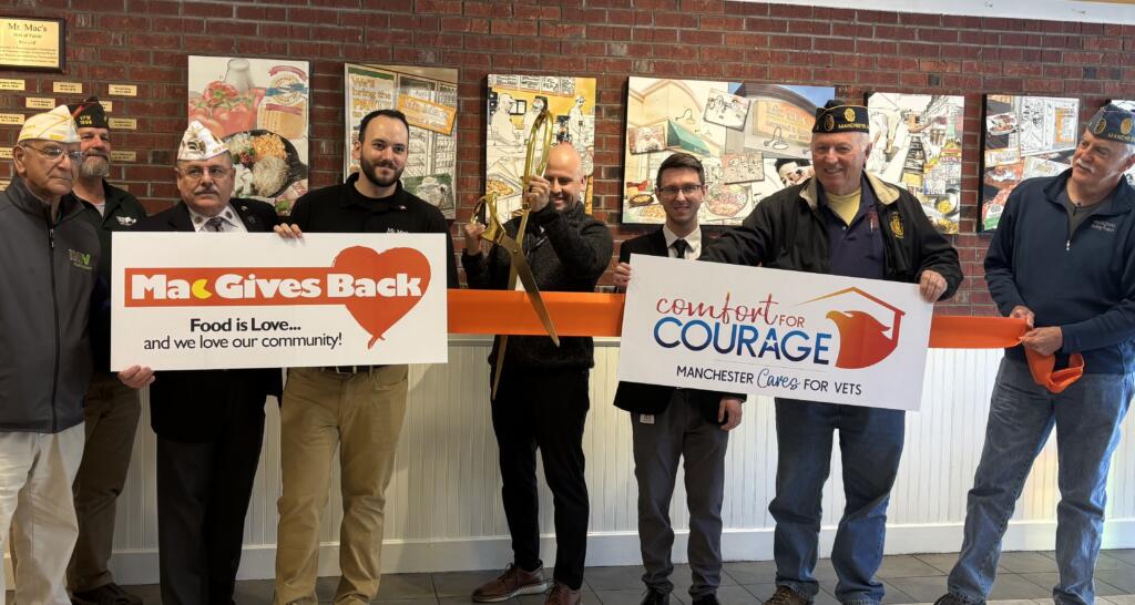 A group of people, some in veterans hats, hold a large ribbon and signs reading Mac Gives Back and Comfort for Courage Manchester Cares for Vets at a ribbon-cutting ceremony in front of a brick wall.