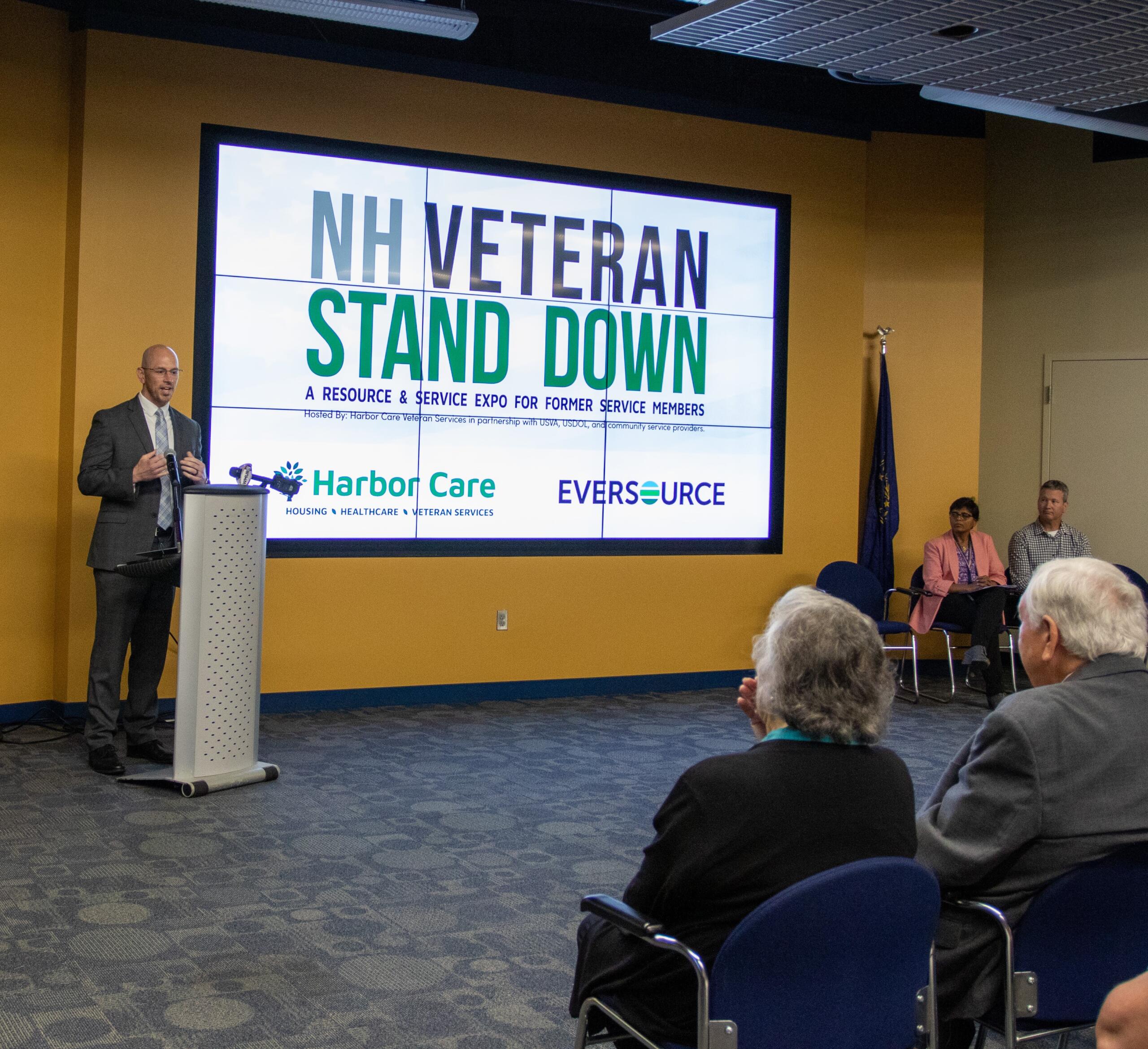 A man speaks at a podium in front of a large screen displaying NH Veteran Stand Down to an audience seated in a conference room.