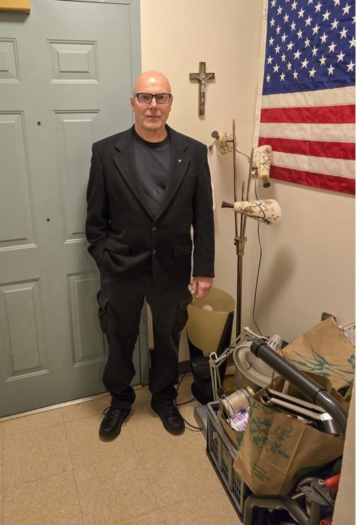 A man wearing glasses, a black suit, and black shoes stands indoors next to a green door. Behind him are a crucifix, an American flag, a lamp, and a box of household items. The floor is tiled.