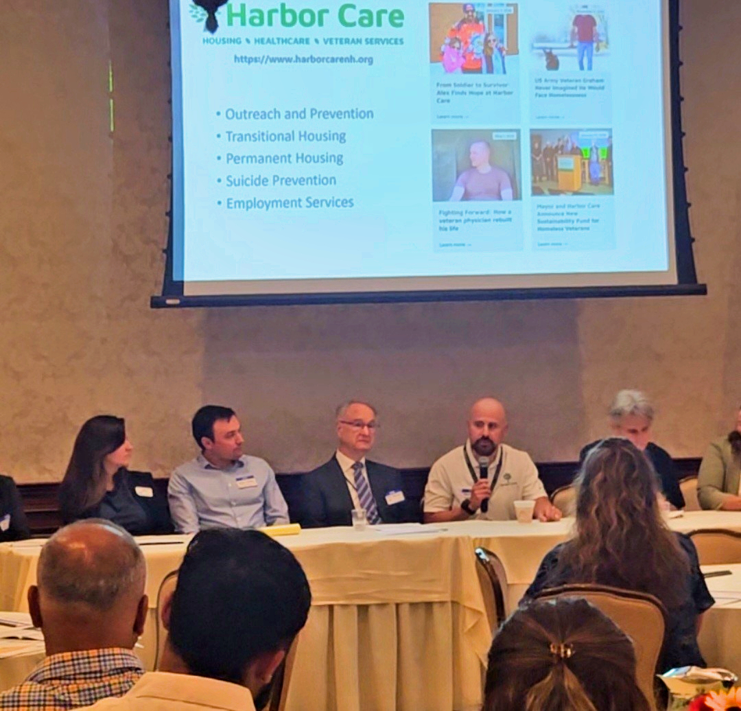 A panel of six people sits at a table speaking to an audience; a presentation slide about Harbor Care services is projected behind them.