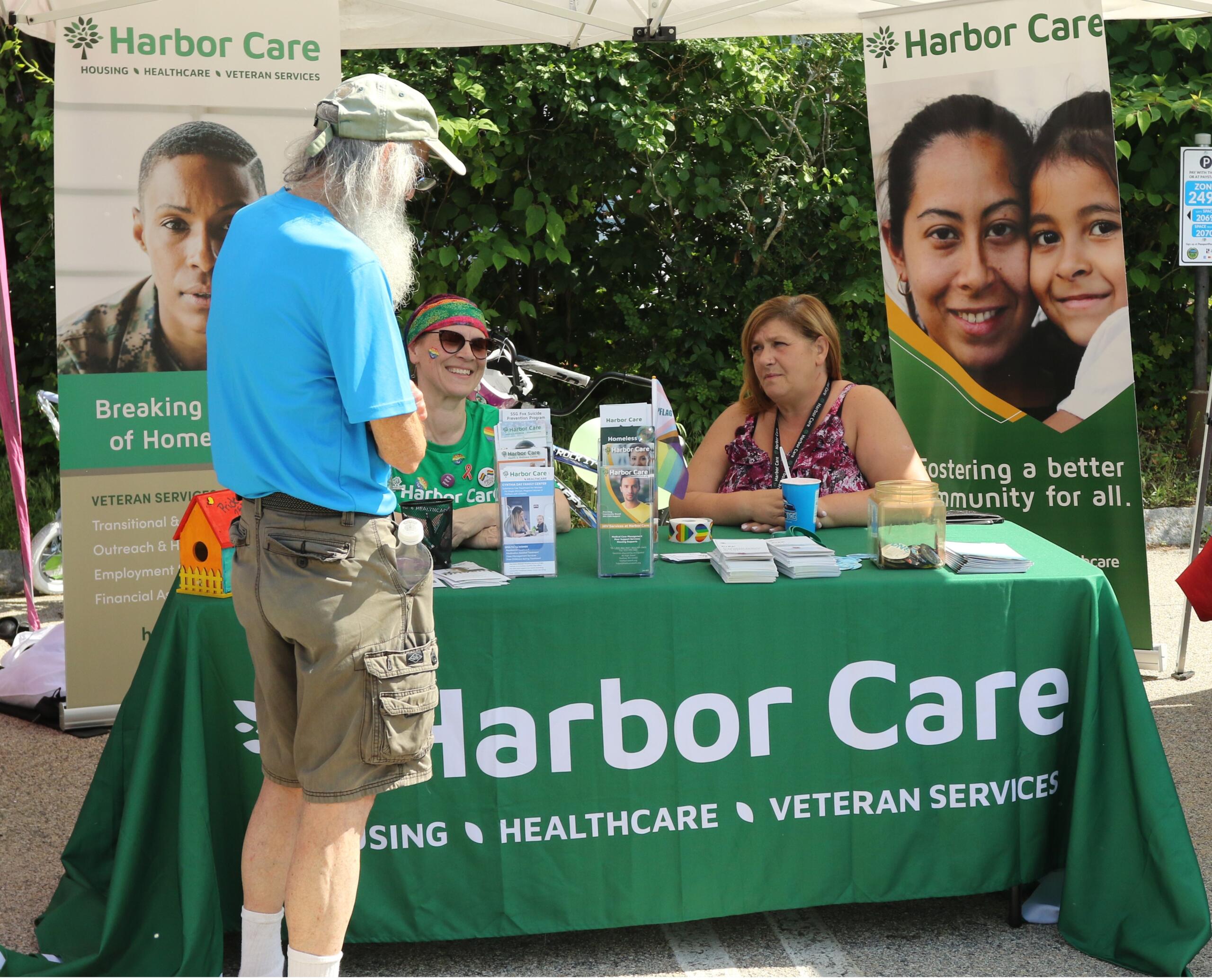5L8A1268 Three people interact at an outdoor Harbor Care booth, which displays information about housing, healthcare, and veteran services. Folded brochures and paperwork are on the table.