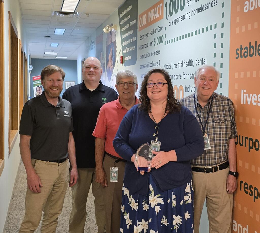 Five adults stand indoors in a hallway with informational wall graphics; the woman in front holds a glass award and smiles at the camera.