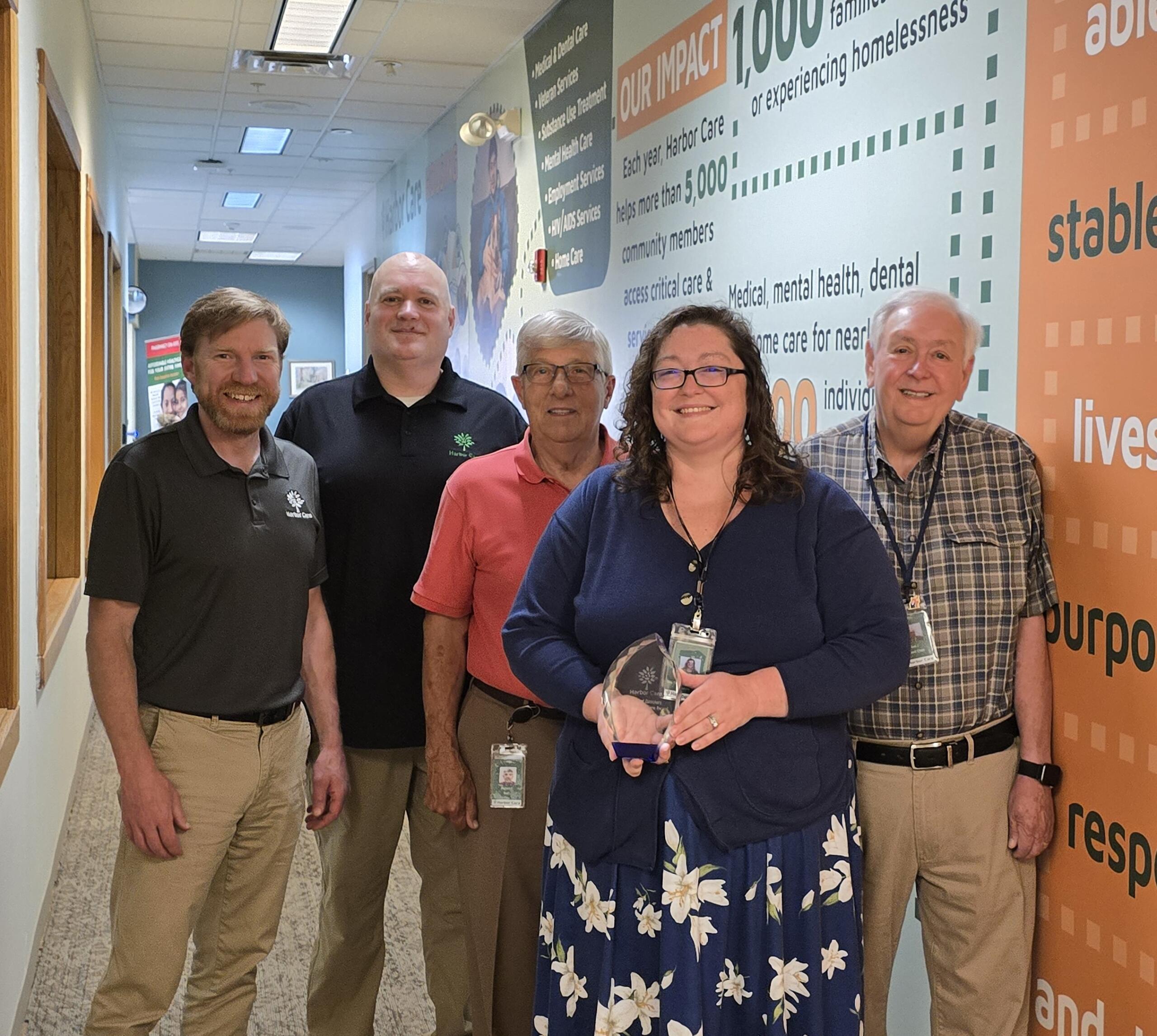 (l-r) Brian Tagliaferro, Chief Philanthropy Officer; Michael Zechman, Human Resources Director; Board Member Rick Plante; Cameron Bergeron, Director of Mental Health Services; and Joel Jaffe, Board Chair. Five adults stand indoors in a hallway with informational wall graphics; the woman in front holds a glass award and smiles at the camera.