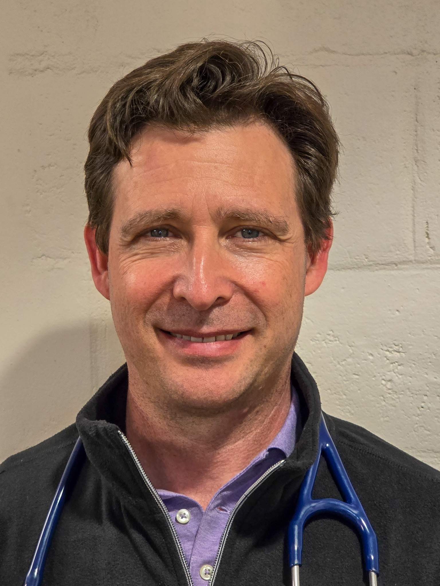 A man with brown hair and blue eyes, wearing a zip-up jacket, collared shirt, and a stethoscope around his neck, stands in front of a light-colored wall.