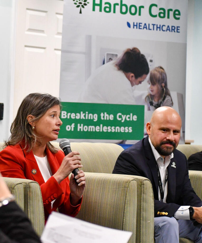 A woman speaks into a microphone while seated next to a man; a Harbor Care Healthcare banner is visible in the background.