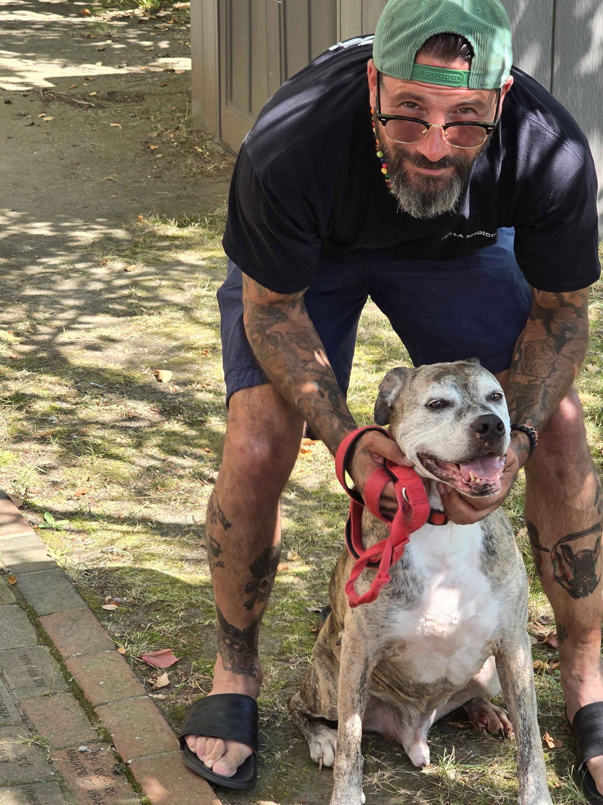 A man with tattoos and glasses, wearing a green cap backwards, holds a smiling brown and white dog with a red leash outdoors on a sunny day.