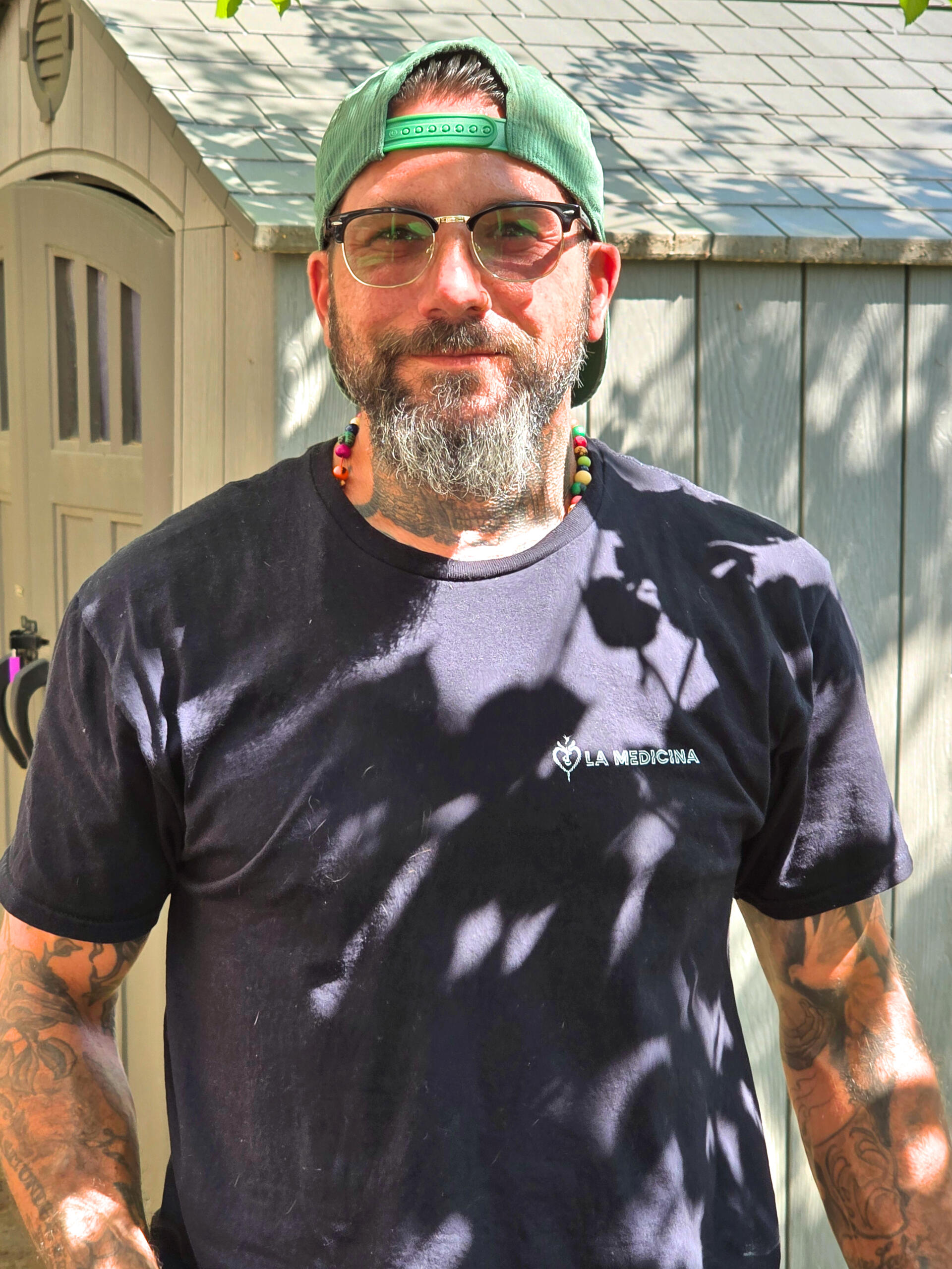 A man with tattoos, glasses, and a backward green cap stands outdoors in sunlight, wearing a navy La Medicina t-shirt and a beaded necklace.