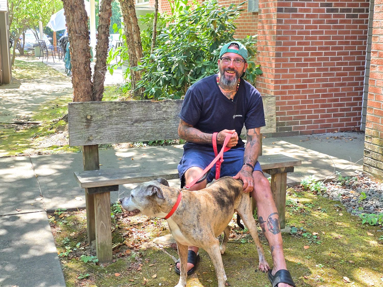 A man sits on a weathered bench outdoors, holding a red leash attached to a brown dog standing in front of him. Trees and a brick building are in the background.