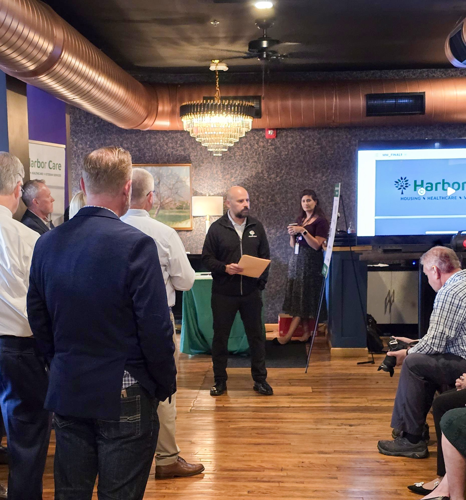 A man speaks to a group of people in a room with wooden floors and a chandelier, while a woman stands near a Harbor Care presentation screen.
