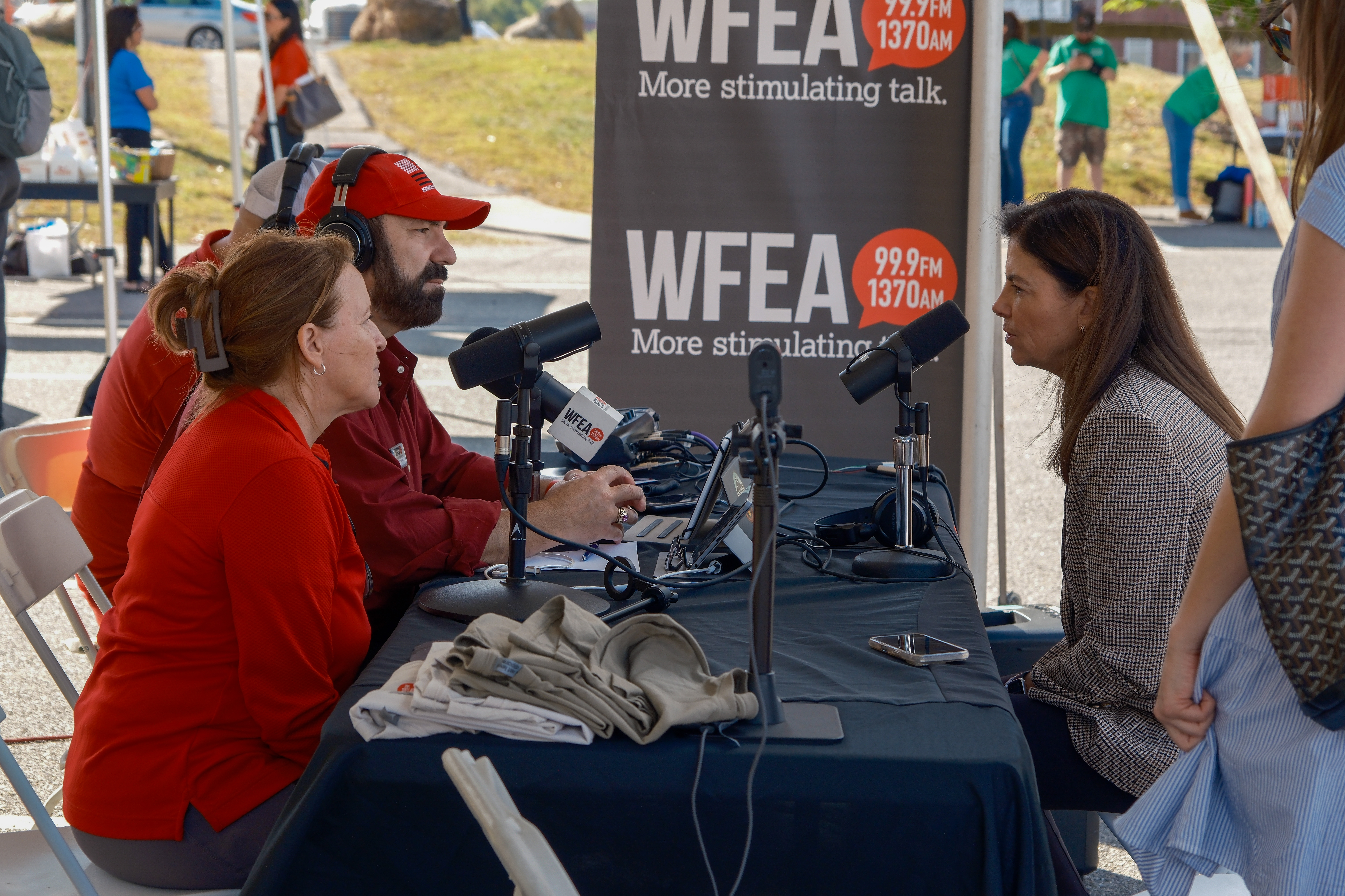 Three people sit at a table with microphones for a live outdoor radio broadcast by WFEA 99.9 FM/1370 AM. One person appears to be interviewed while others operate equipment.