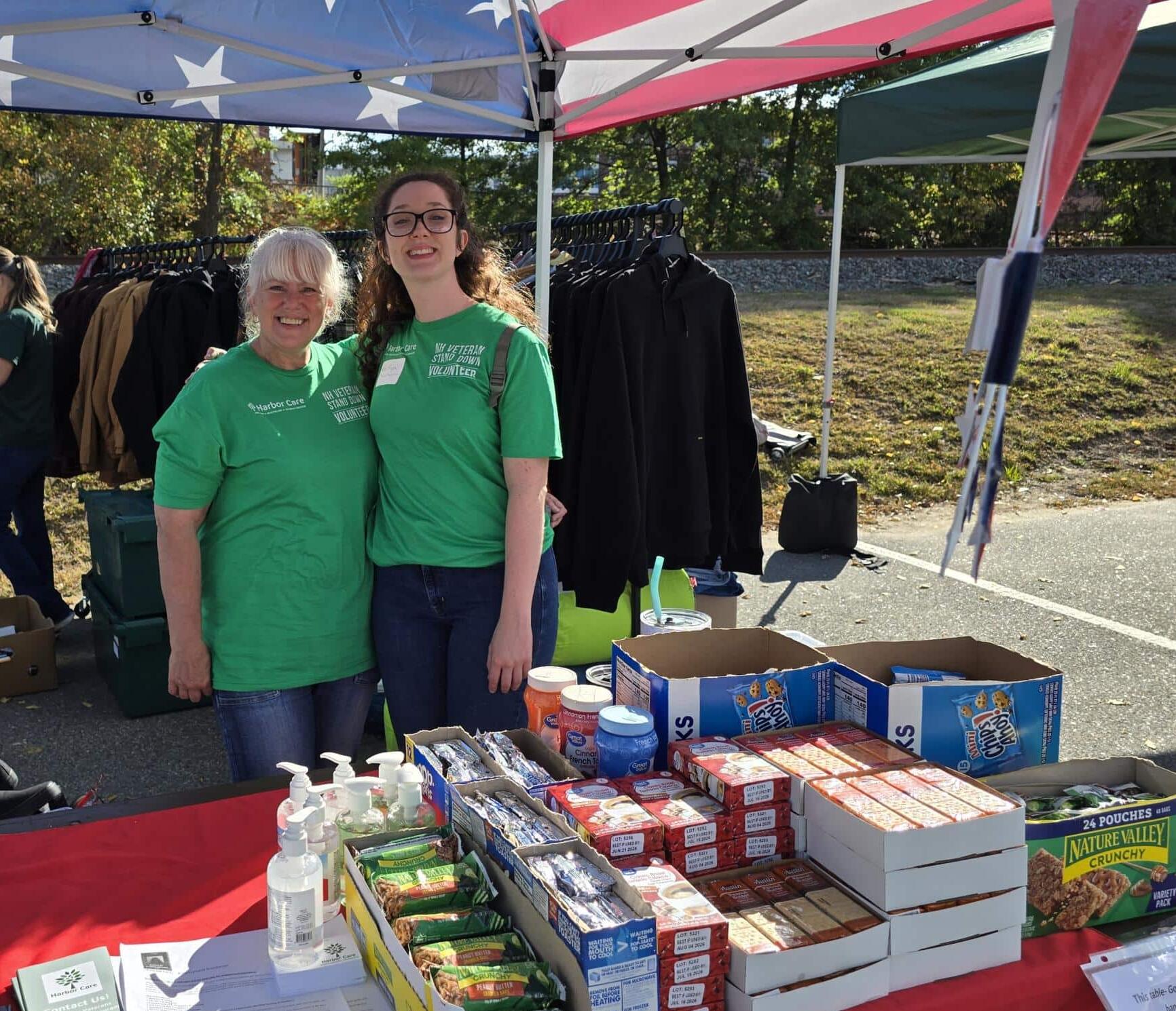 Two women in green shirts stand under a tent with an American flag canopy, behind a table displaying snacks, sanitizer bottles, and informational materials at an outdoor event.