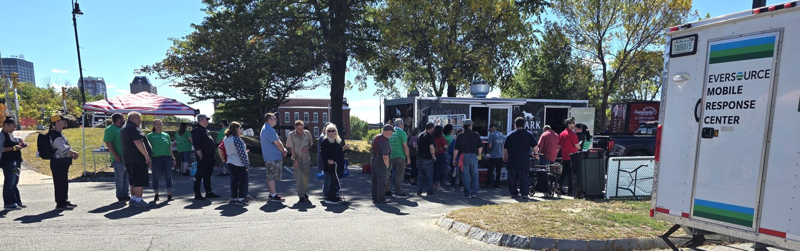 20250919_124025 People stand in a line outdoors near food trucks and an Eversource Mobile Response Center vehicle on a sunny day with trees and tents in the background.