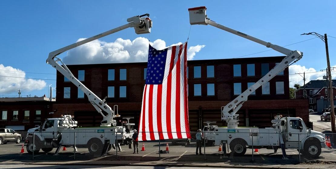 548275885_1102934828700613_3205549648043761765_n (1) Two utility trucks with raised buckets hold a large American flag between them in front of a brick building on a sunny day.
