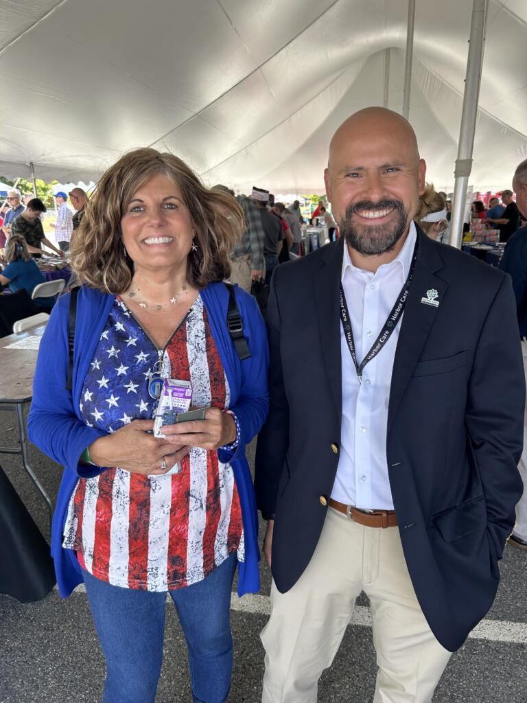 Two people stand under a large event tent. The woman wears an American flag shirt and blue cardigan; the man wears a blazer and lanyard. Both are smiling at the camera.