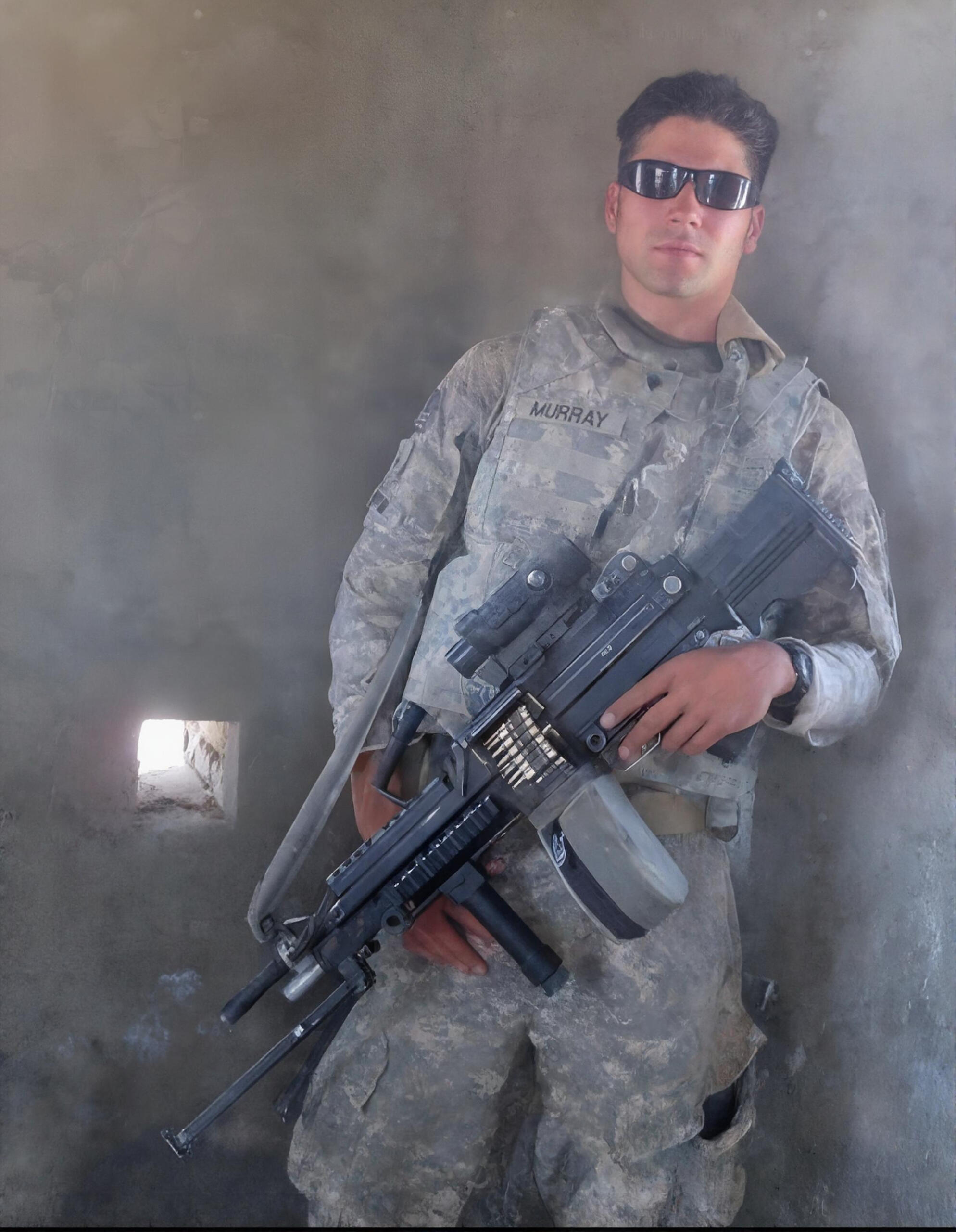 A soldier in camouflage uniform and sunglasses stands indoors, holding a machine gun. The name MURRAY is visible on his uniform. Light enters through a small hole in the wall.