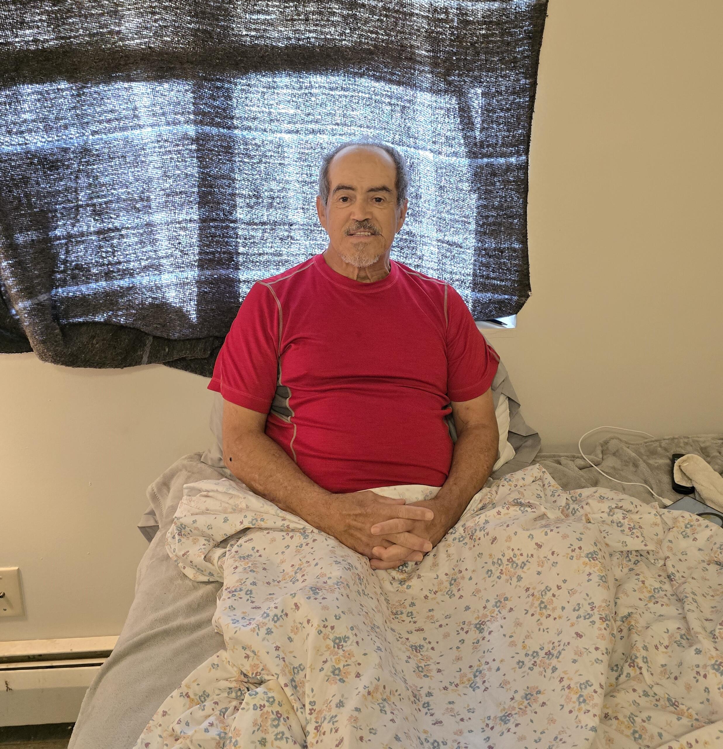 A middle-aged man with short hair and a mustache sits upright in bed, wearing a red shirt, with a patterned blanket over his legs. A dark curtain covers the window behind him.