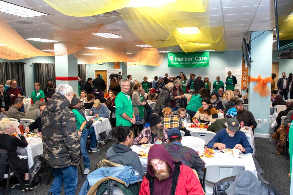 A large group of people sits and stands in a decorated community hall, eating and socializing at tables under orange and yellow streamers. A green Harbor Care banner hangs on the wall.