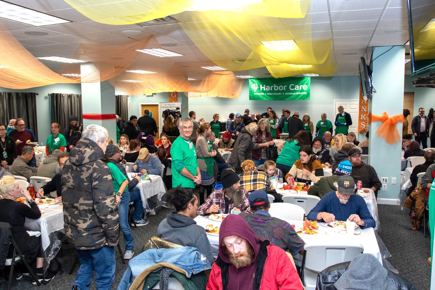A large group of people sits and stands in a decorated community hall, eating and socializing at tables under orange and yellow streamers. A green Harbor Care banner hangs on the wall.