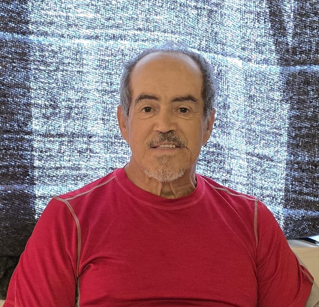 An older man with short gray hair and a trimmed beard, wearing a red shirt, sits indoors in front of a window covered with a dark patterned curtain.