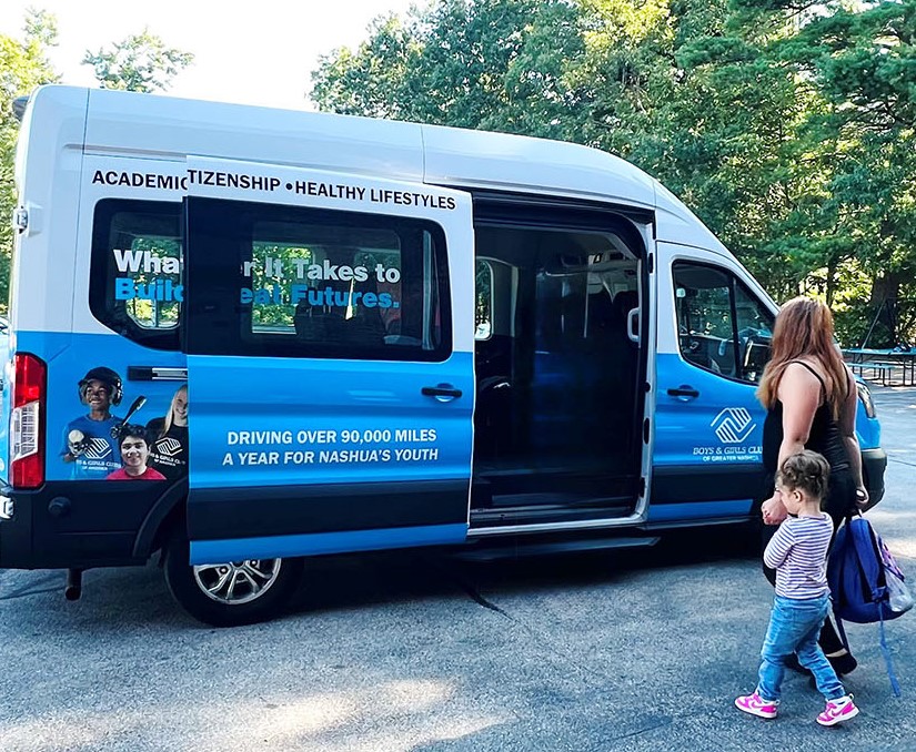 A woman and a young girl walk toward a blue Boys & Girls Club van parked on a paved surface, with trees in the background.