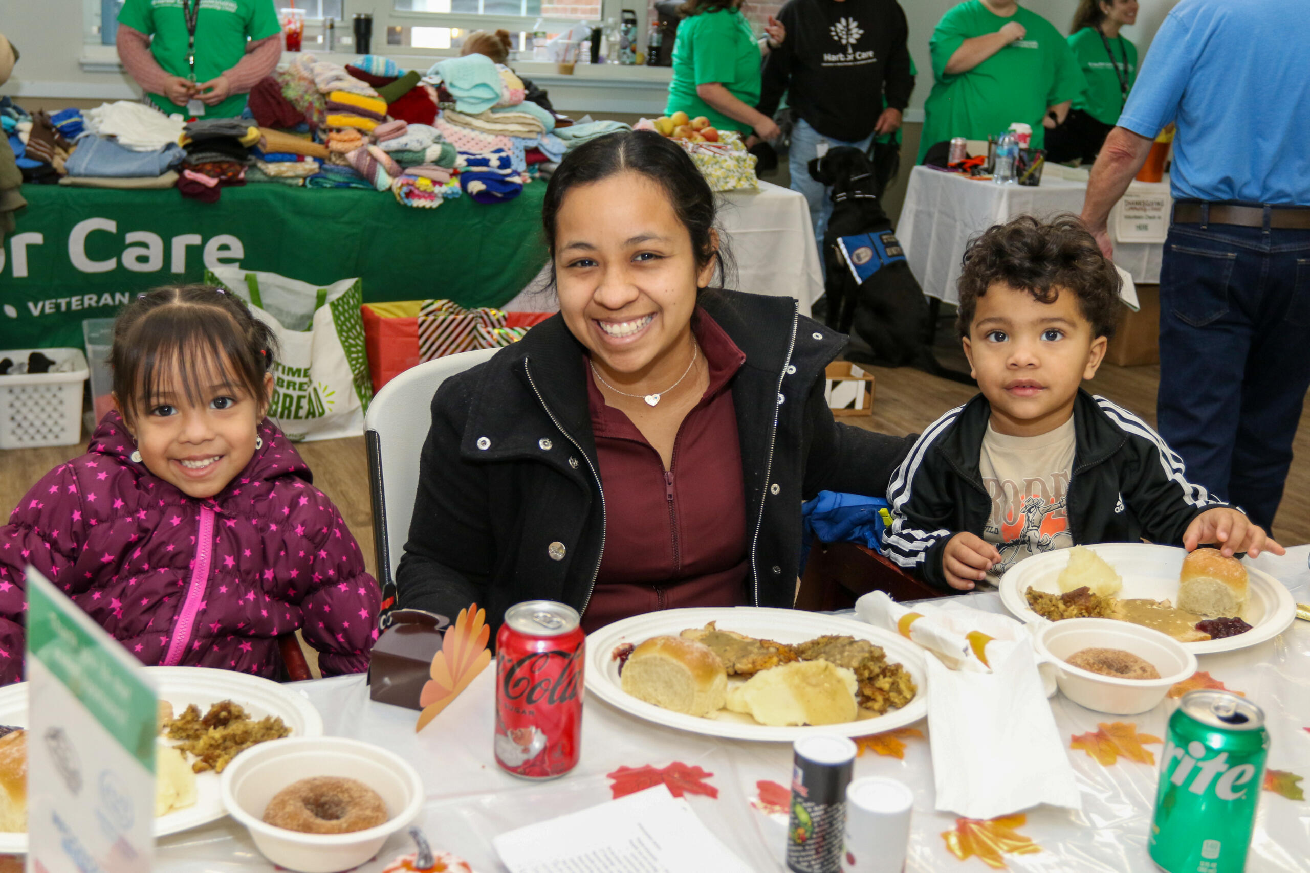 A woman and two children are seated at a table with plates of food and drinks, smiling at the camera. People and tables with items are in the background.