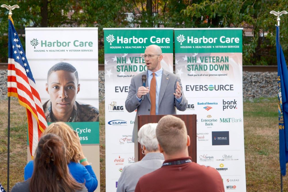 A man speaks at an outdoor event in front of Harbor Care banners and an American flag, with attendees seated and trees in the background.