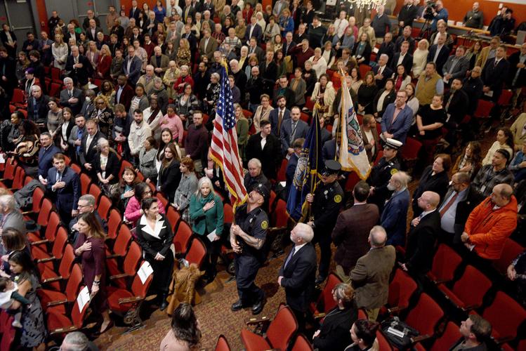 A large crowd stands in an auditorium, many with hands on their hearts, as uniformed personnel carry flags down the aisle.