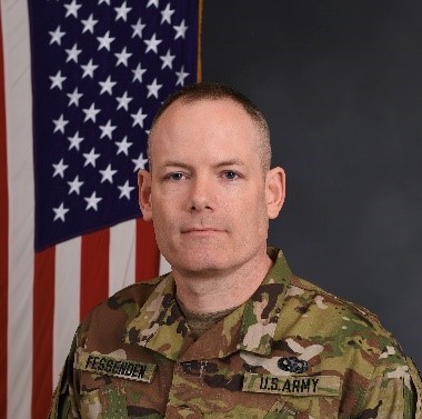 A man in U.S. Army camouflage uniform poses in front of an American flag on a dark background.