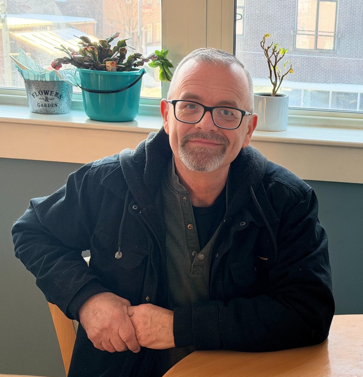 IMG_0017-fotor-20260121192319 A man with glasses and a beard sits at a table indoors, smiling slightly. Potted plants are on the windowsill behind him.