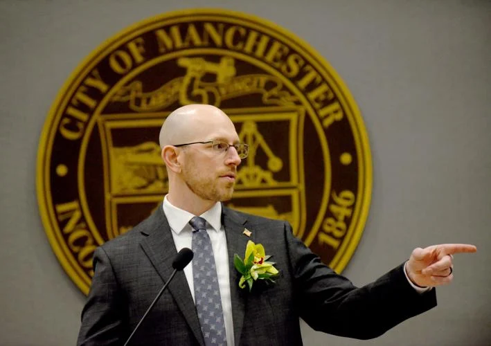 A man in a suit with a flower boutonniere speaks and gestures while standing in front of a City of Manchester seal.