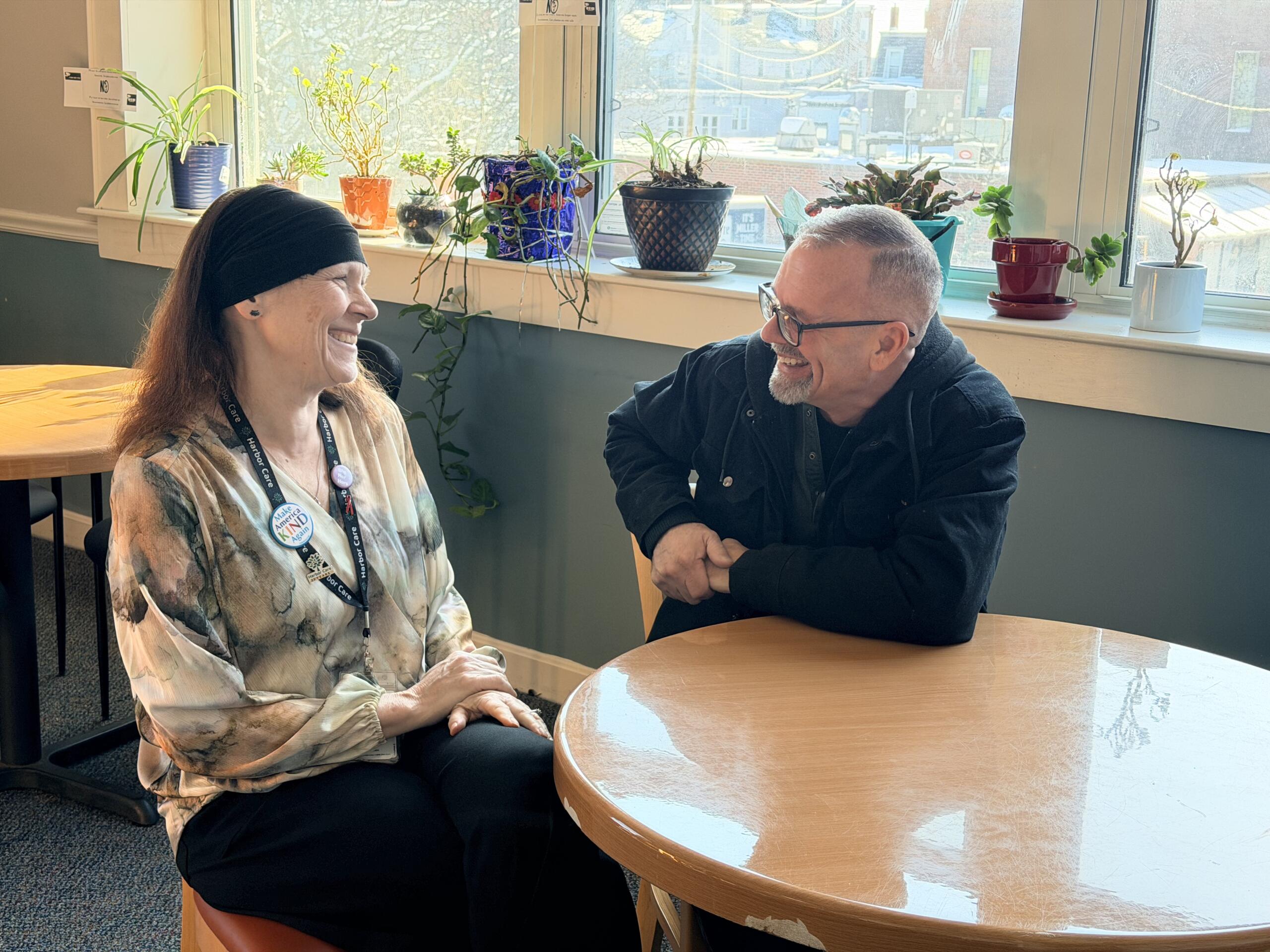 Two people sit at a round table smiling and talking in a sunlit room with potted plants on the windowsill behind them.
