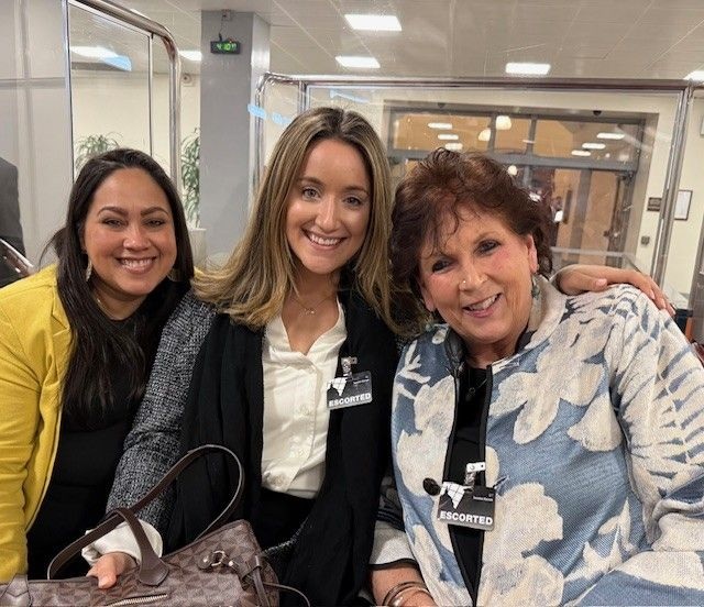 (l-r): Olga Rodriguez, Harbor Care Health & Wellness Center Practice Administrator; Jessica Campbell, recipient and Manager of Staff Development; and Tess Kuenning, President & CEO of Bi-State Primary Care Association. Three women sitting closely together indoors, all smiling at the camera. Two wear ESCORTED badges, and one holds a handbag.