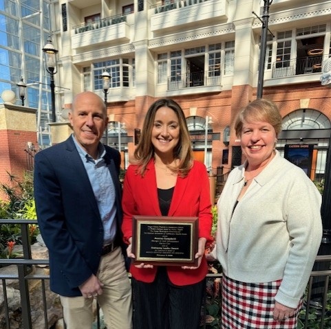 William Belecz, Harbor Care Chief Operating Officer; Jessica Campbell, recipient and Manager of Staff Development; and Board Member Rosemarie Dykeman. Three people stand together indoors; the person in the middle holds a rectangular plaque, while the group smiles at the camera. The background features large windows and a multistory building interior.