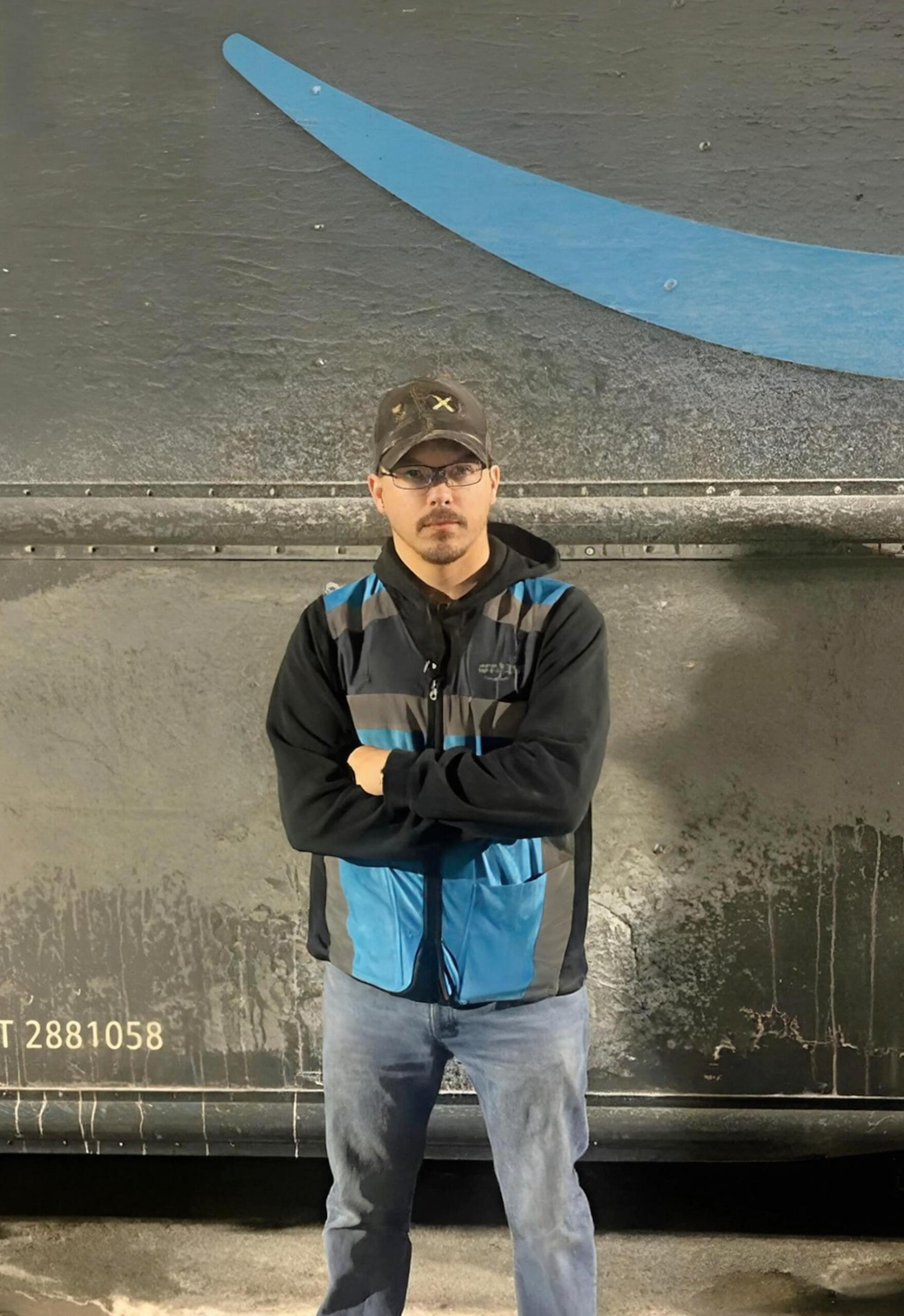 A man wearing glasses, a cap, and a blue-and-black jacket stands with his arms crossed in front of a dark, dirty truck with a partial blue logo.