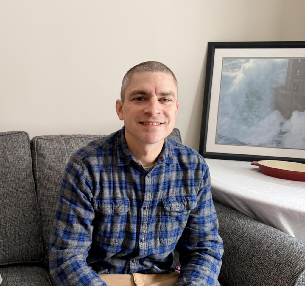 A man wearing a blue plaid shirt sits on a gray couch. Behind him is a framed ocean photograph and a white table with a red-edged dish.