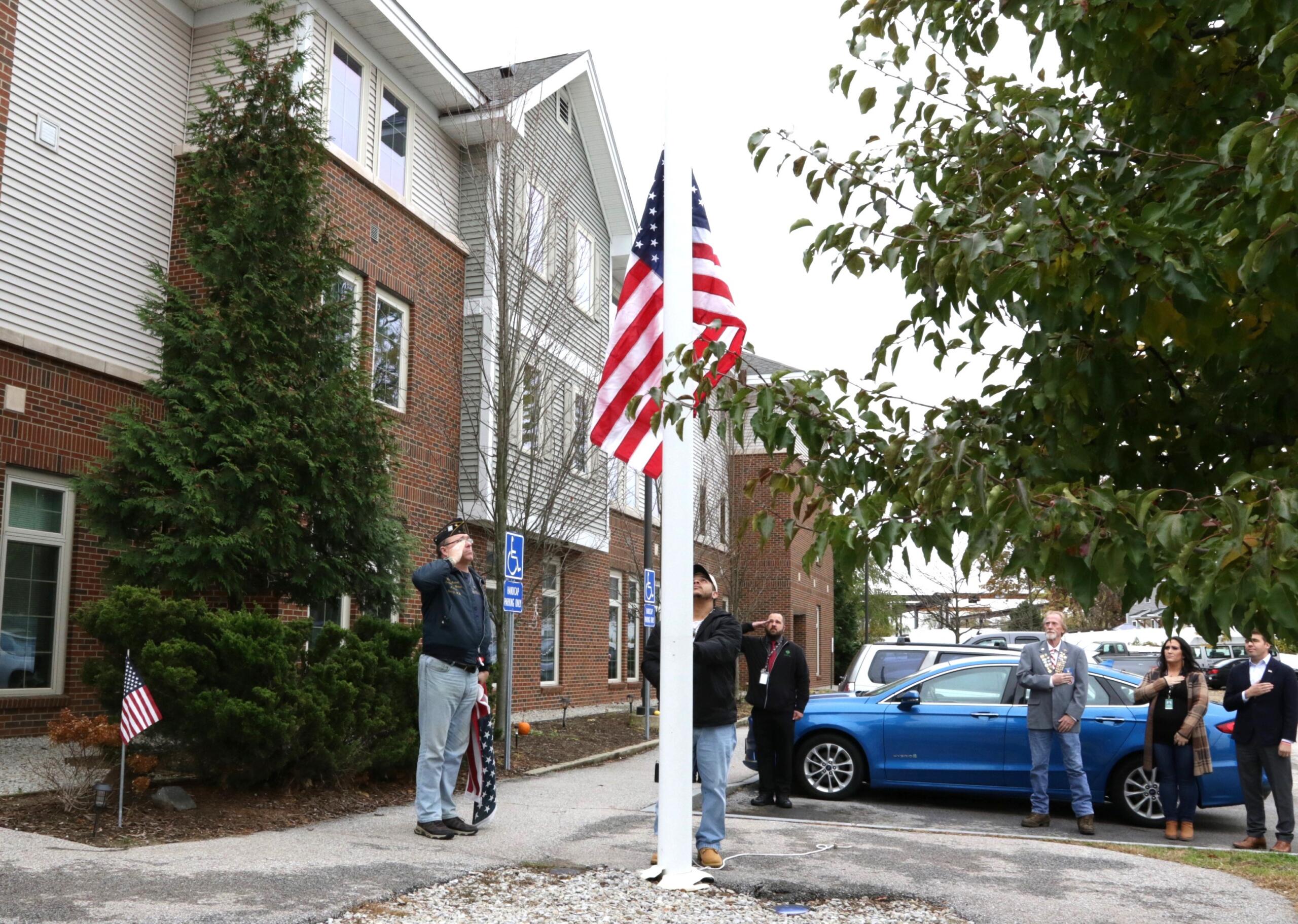 People stand and salute as the American flag is raised on a flagpole outside a brick building, with parked cars and trees nearby.