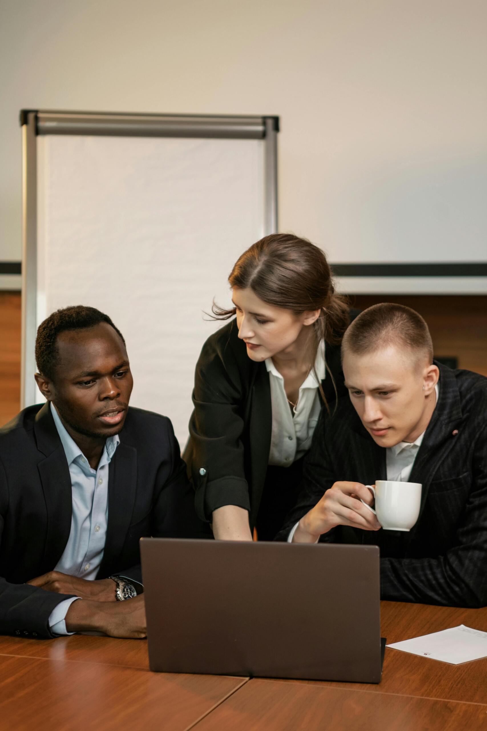Three people in business attire are gathered around a laptop at a meeting table, with a blank flip chart in the background. One person is holding a white mug.