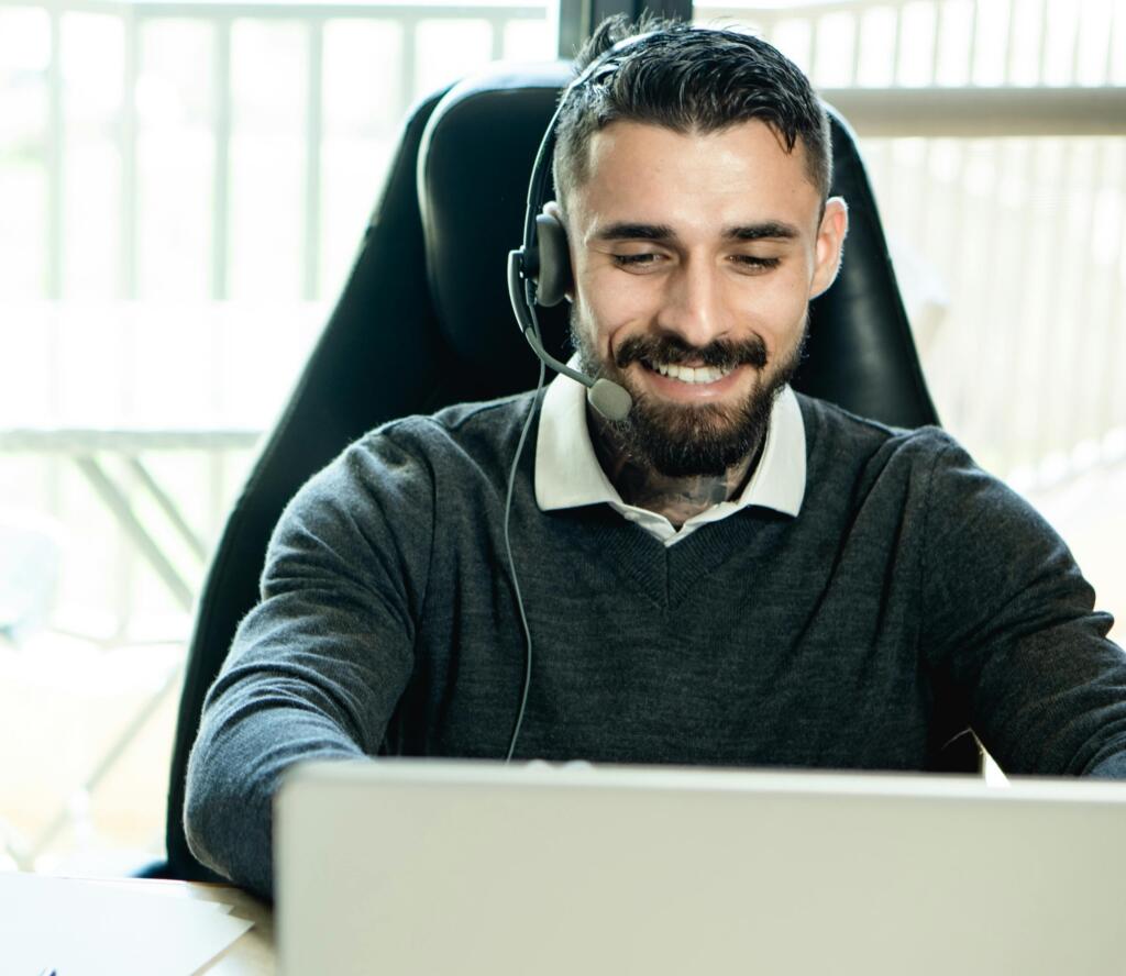 A man with a beard wearing a headset and a sweater sits at a desk, smiling while looking at a laptop.