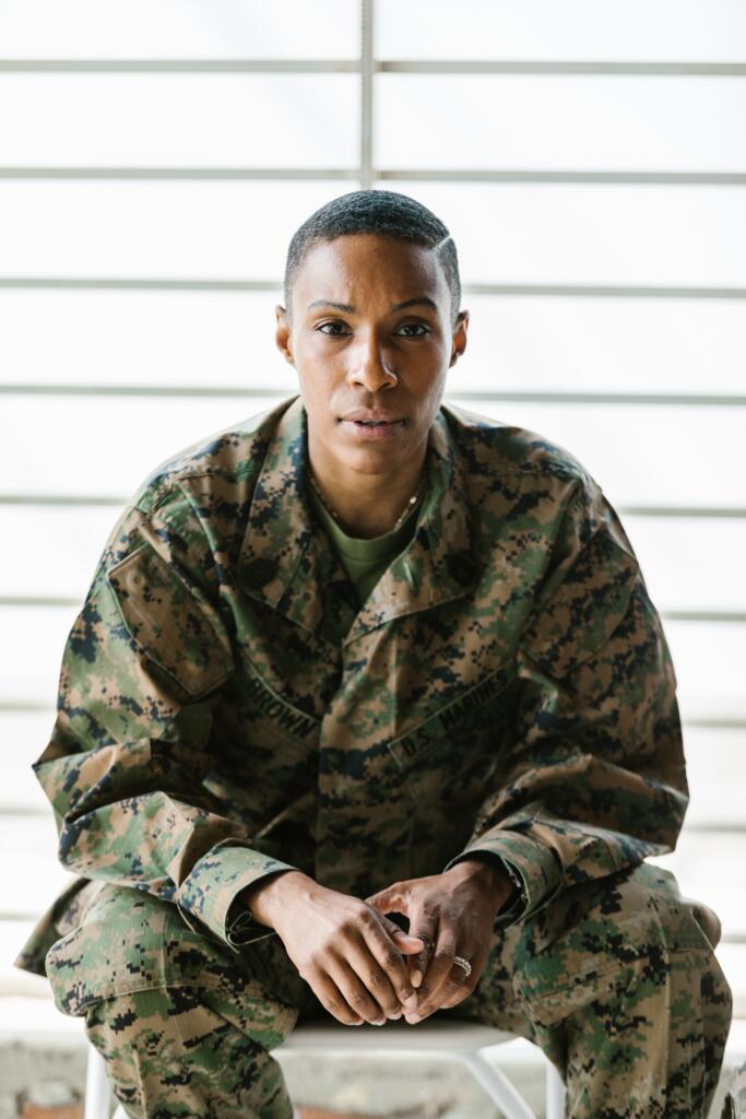 A person in military camouflage uniform sits on a stool, facing forward with hands resting together, in front of a bright, slatted background.