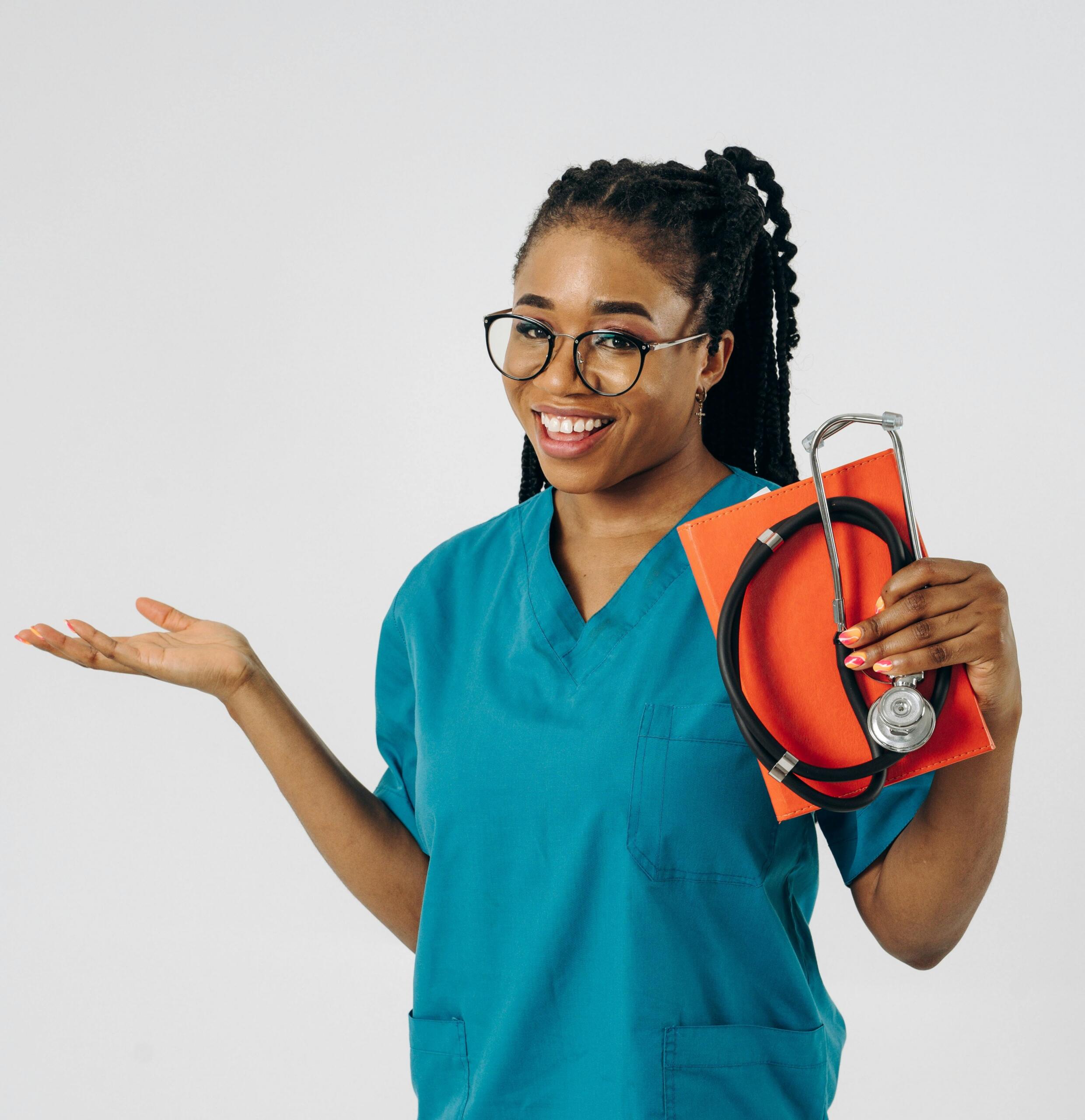 Smiling healthcare worker in teal scrubs holds a stethoscope and orange notebook, gesturing with one hand against a plain white background.