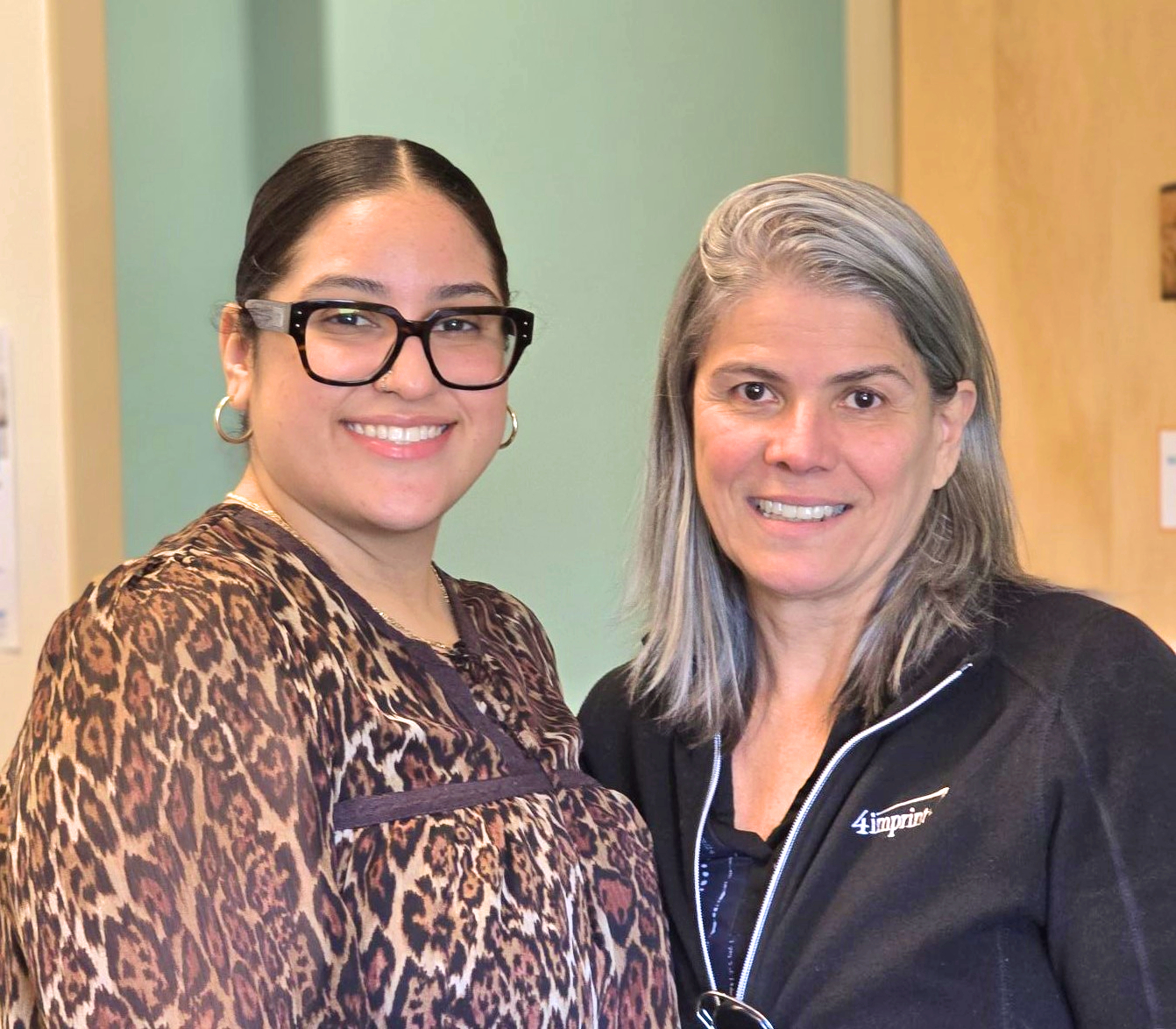 Two women standing side by side indoors, smiling at the camera; one wears glasses and a leopard print top, the other has gray hair and a black jacket.