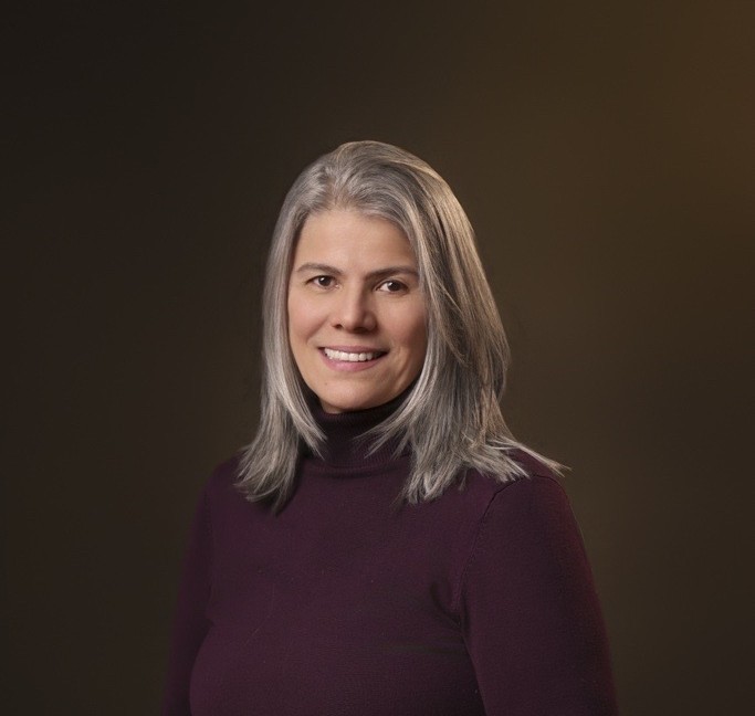 Woman with straight gray hair wearing a dark purple turtleneck, smiling, posed against a plain brown background.
